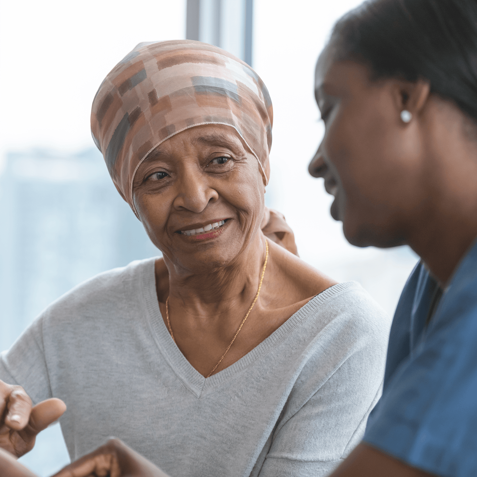 Older Black woman in patterned head scarf smiles as she listens to a younger Black woman in blue scrubs.